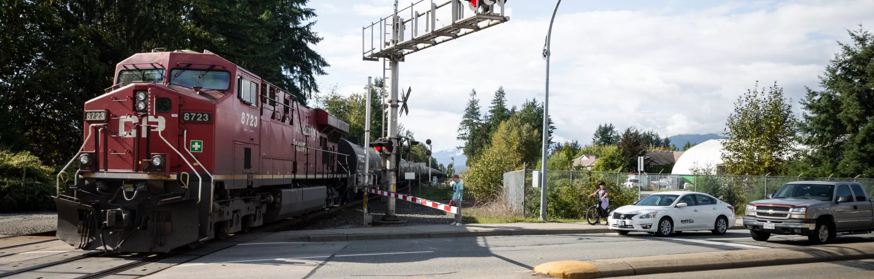 A red CP train can be seen crossing a road on the left. One the right, cars and pedestrians wait at the rail crossing.