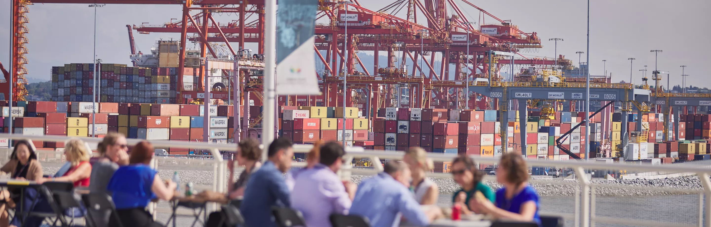 Des employés de l'autorité portuaire sont assis à des tables de pique-nique en plein air. Des grues à portique orange et des conteneurs d'expédition sont visibles à l'arrière-plan.