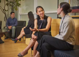 Two women sit in the foreground on a wooden bench in an office talking. In the background, a man and women sit on dark armchairs talking.