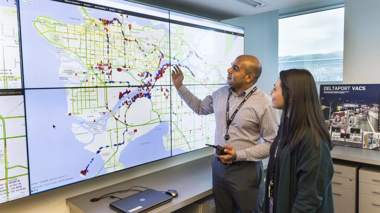 A man and a woman stand in front of a wall of monitors displaying a GPS map.